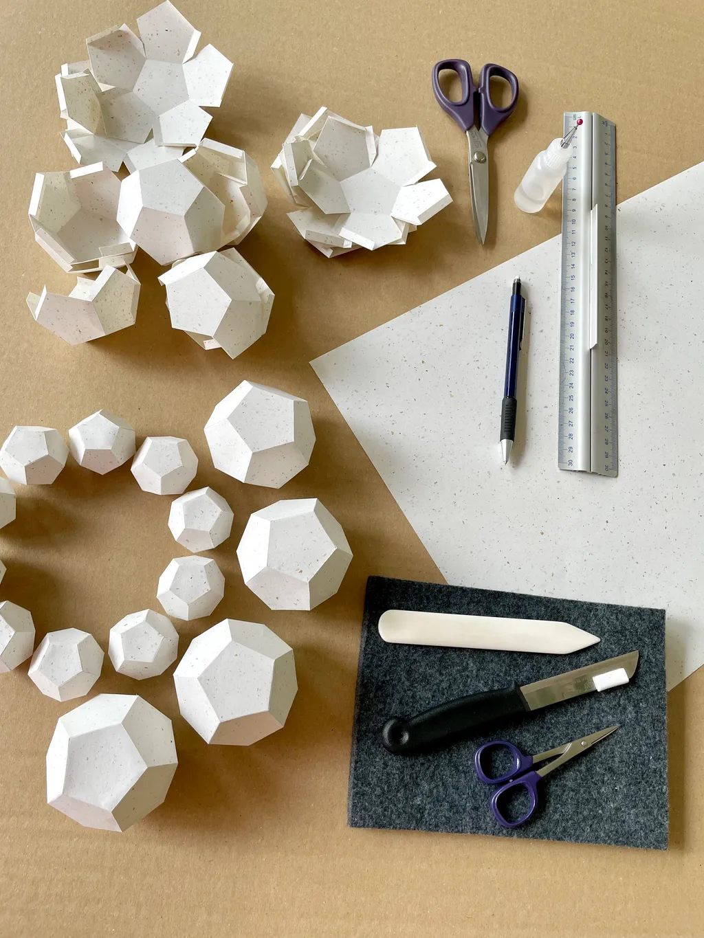 Big Art Show 2026 – Folded polyhedra, a sheet of paper, bone folder, knife, scissors and ruler on the worktable