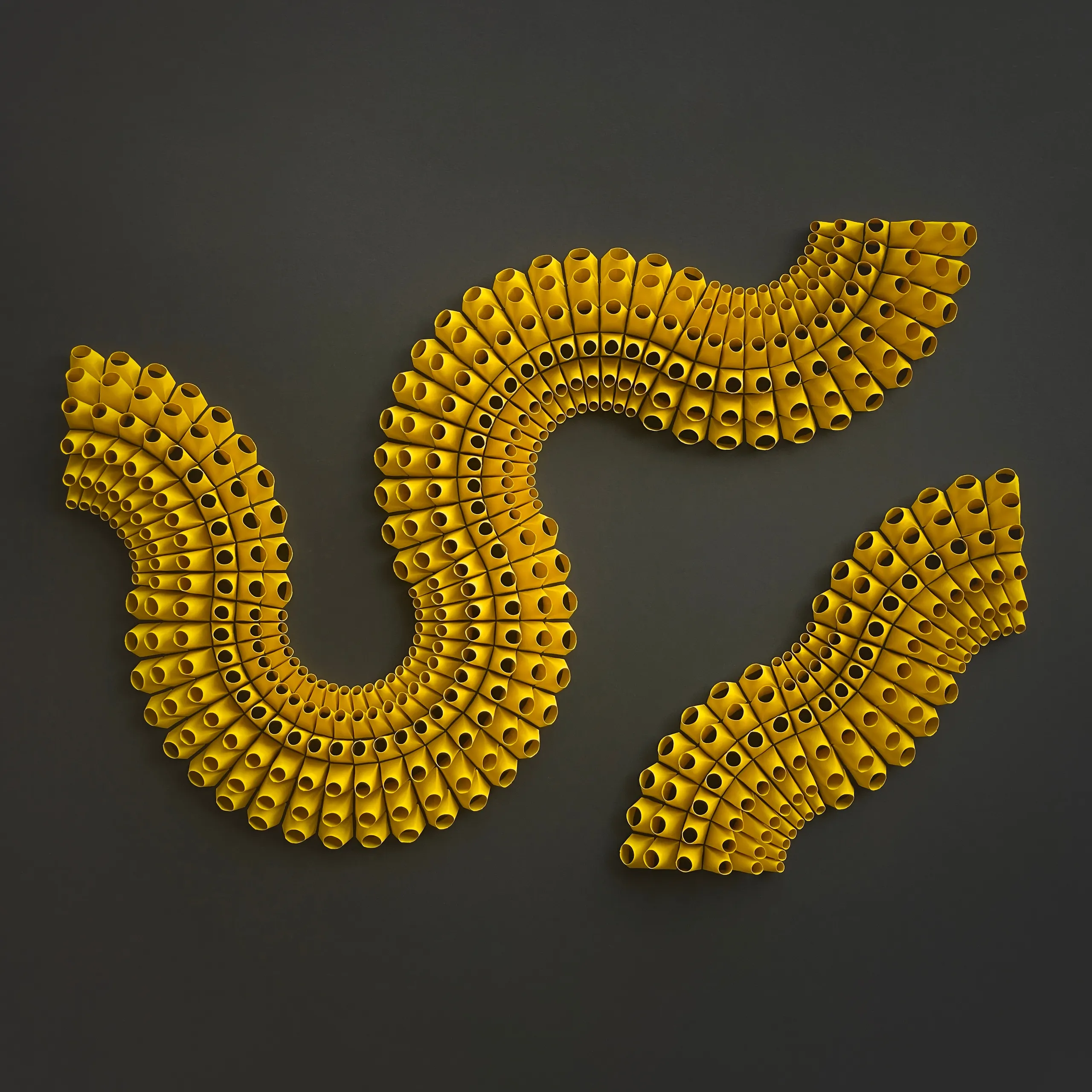 The whole work head-on, three yellow bands of open paper tubes winding across a dark grey ground